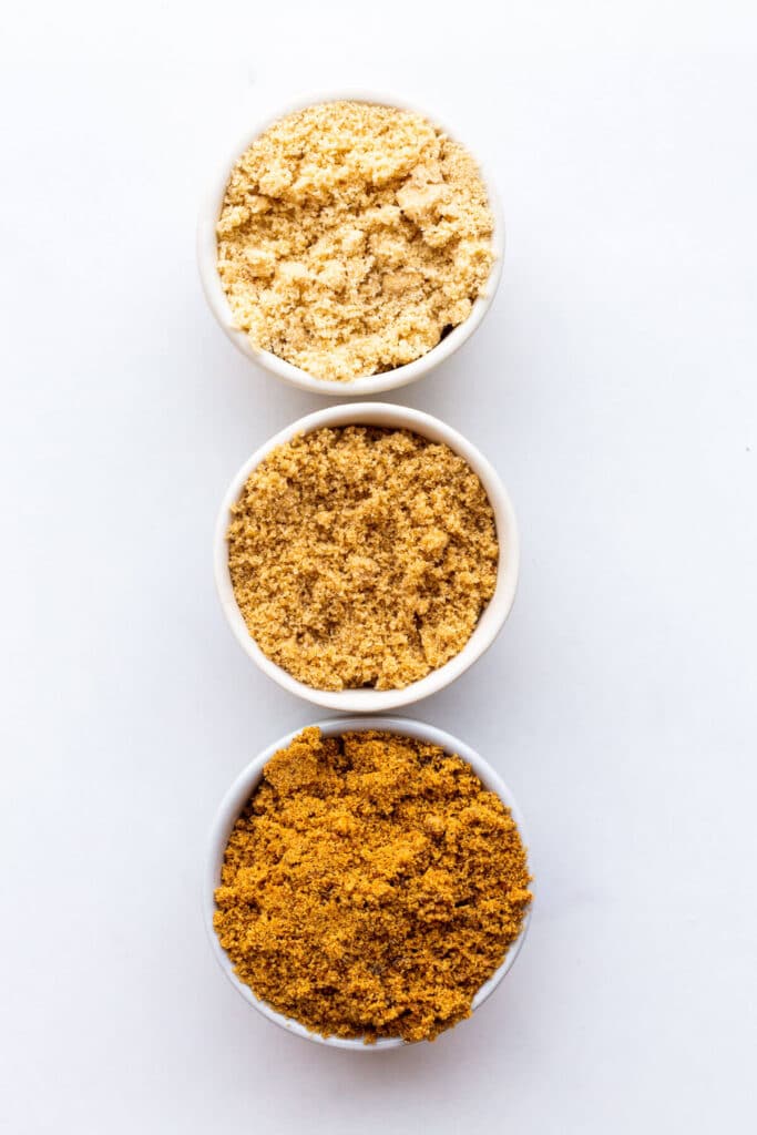 Three bowls of brown sugar to show the difference between light brown sugar (lightest in colour), dark brown sugar (medium in colour), and muscovado sugar (the darkest in colour).