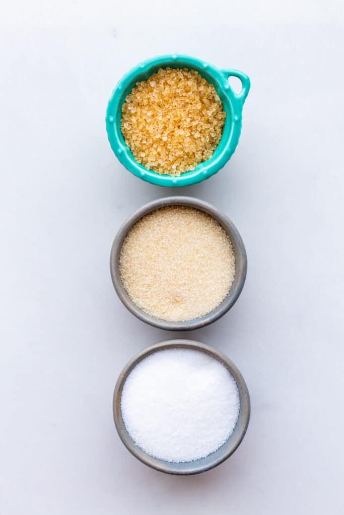 Three bowls of sugars to show the difference between turbinado sugar (also called demerara sugar) with golden medium crystals, cane sugar (golden fine crystals), and granulated sugar (fine white crystals).