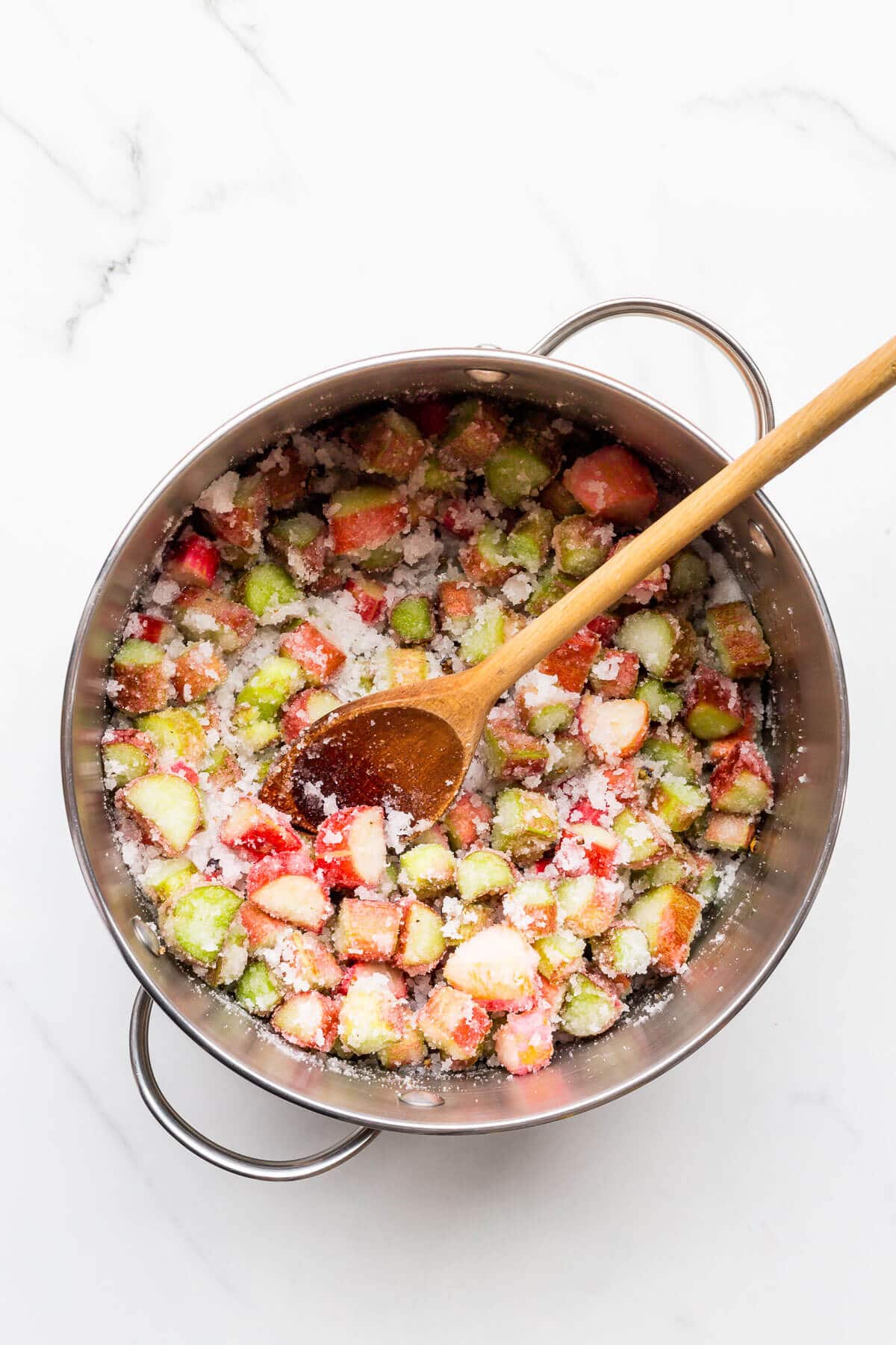 A pot of chopped rhubarb jam mixed with sugar before boiling to the jam setting point, stirred with a wooden spoon.