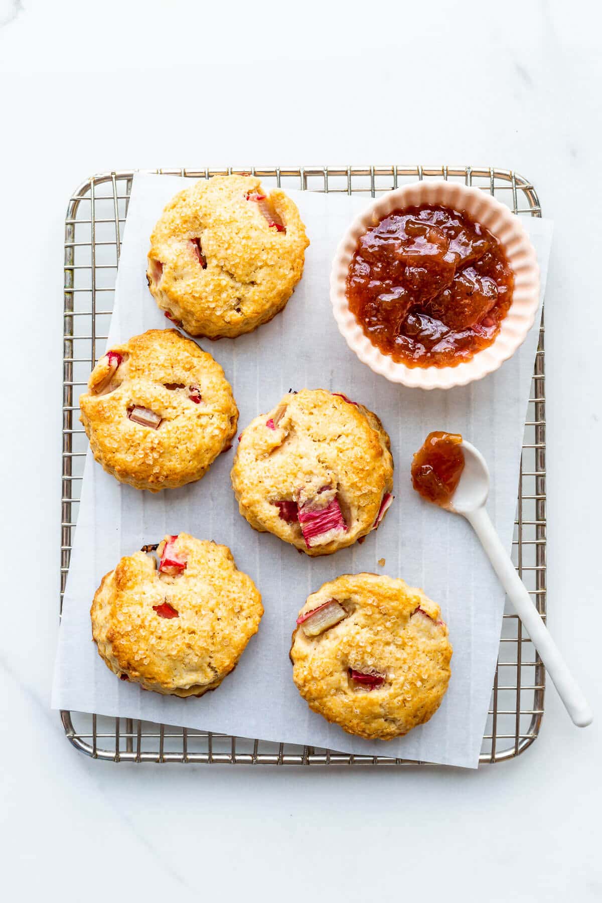 Freshly baked scones with rhubarb on a small cooling rack with a bowl of jam.