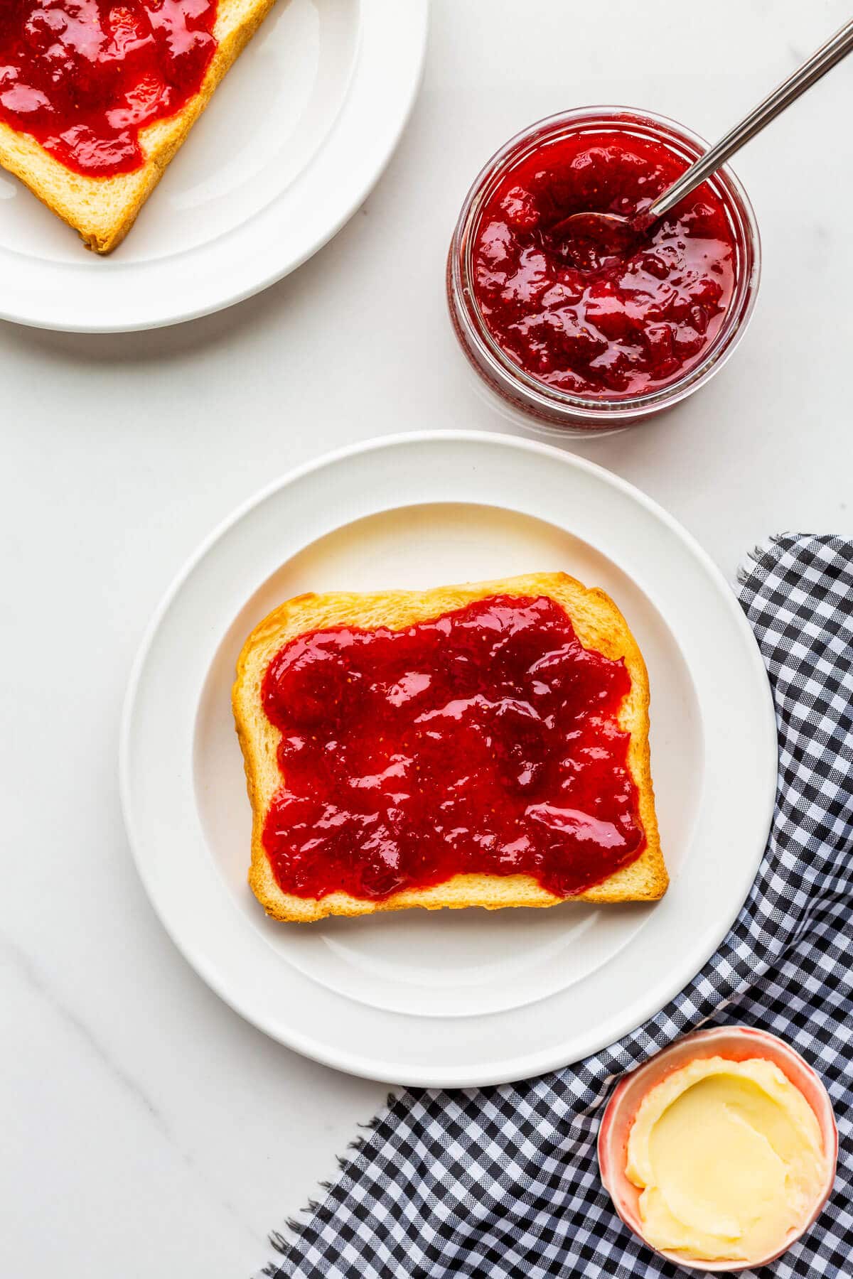 A plate with a piece of toast smeared with strawberry rhubarb jam and served with butter on the side.