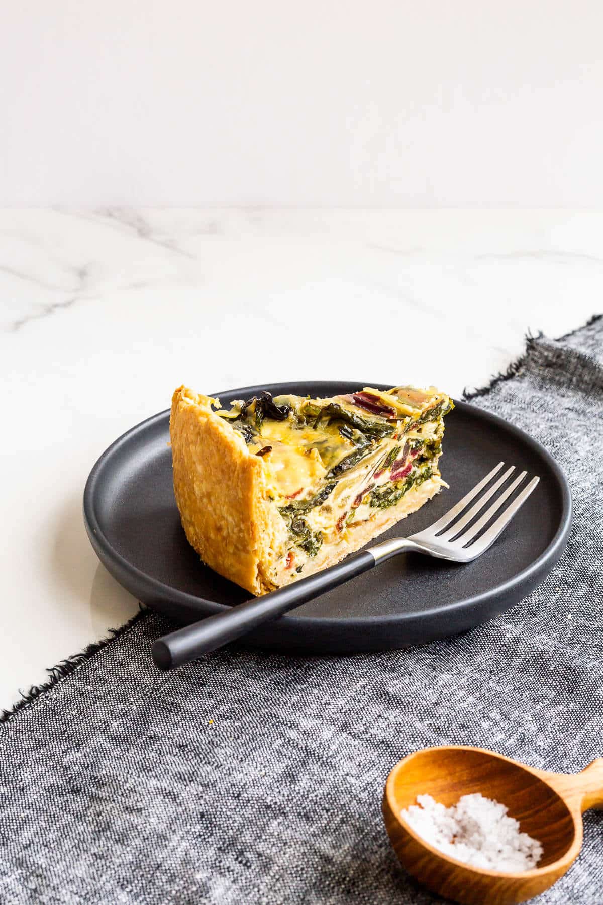 A slice of quiche with rainbow Swiss chard in it, served with a small bowl of salt on the side.