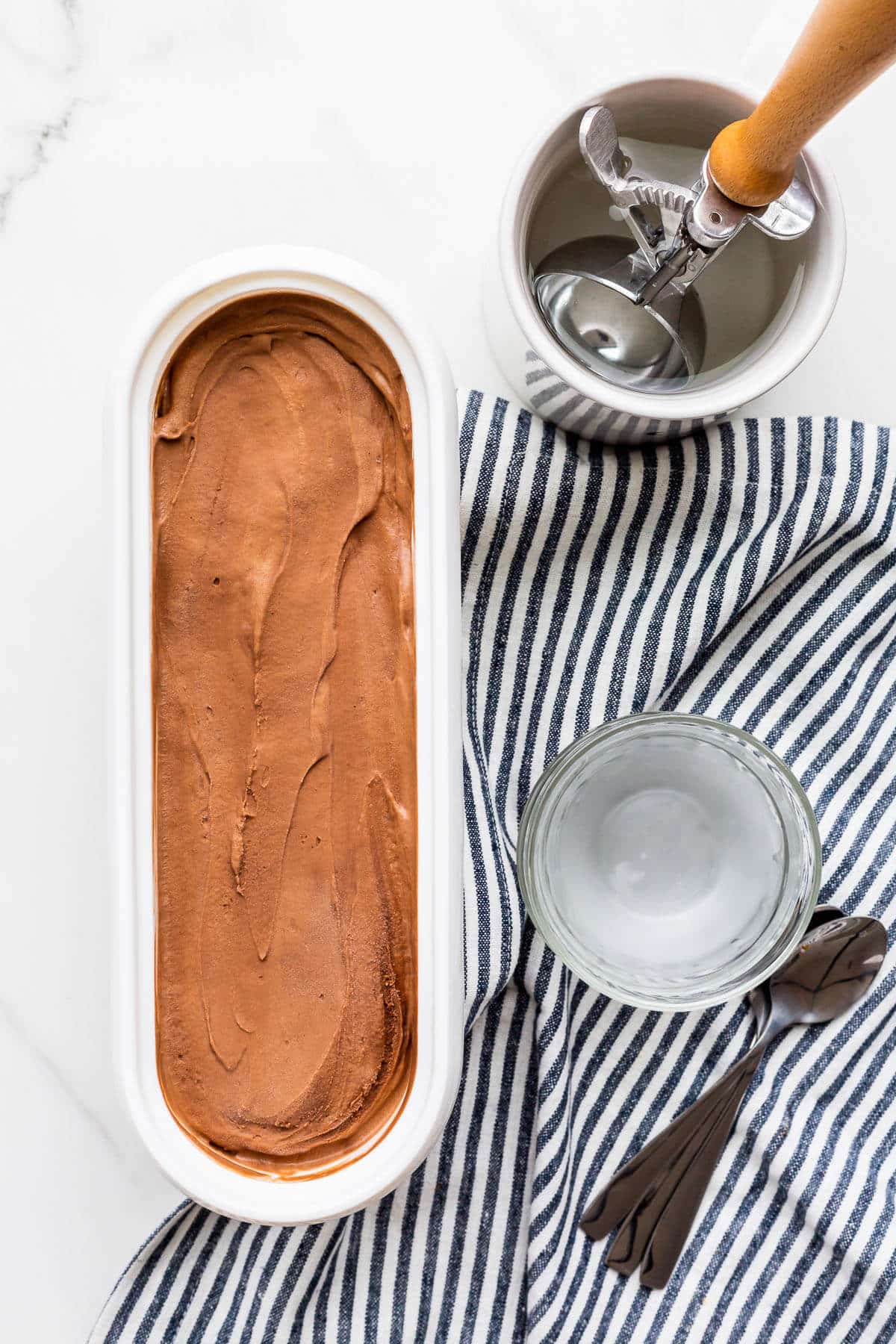 A tub of chocolate ice cream ready to be scooped with an ice cream scoop and served in glass bowls.