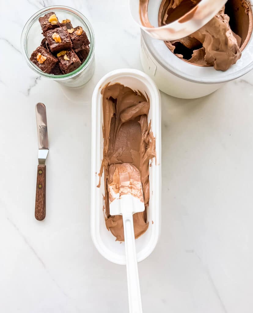 Spreading a thin layer of freshly churned chocolate ice cream at the bottom of a tub to layer with brownie pieces.
