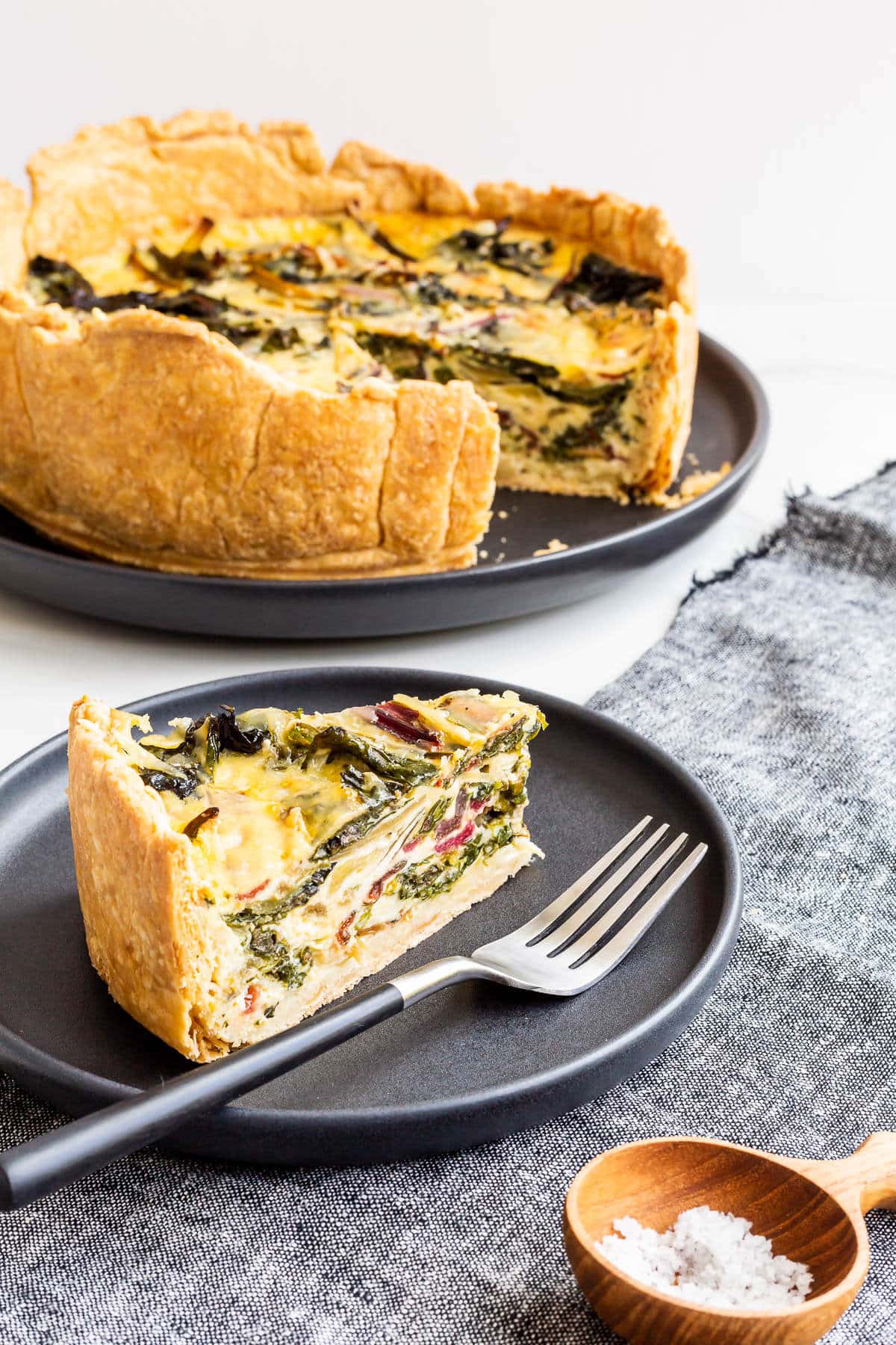 A slice of quiche on a black plate and in the background the deep dish quiche with the slice removed to show the inside with colourful rainbow Swiss chard.