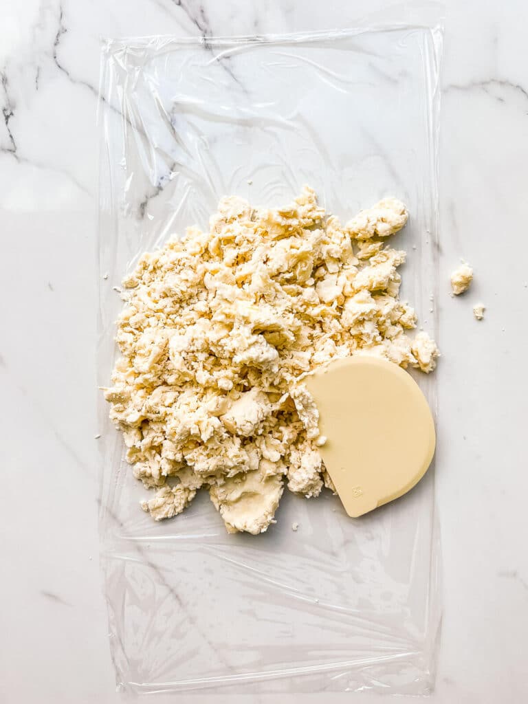 Pressing pie dough together to shape into a disk with a bench scraper on a piece of plastic wrap.