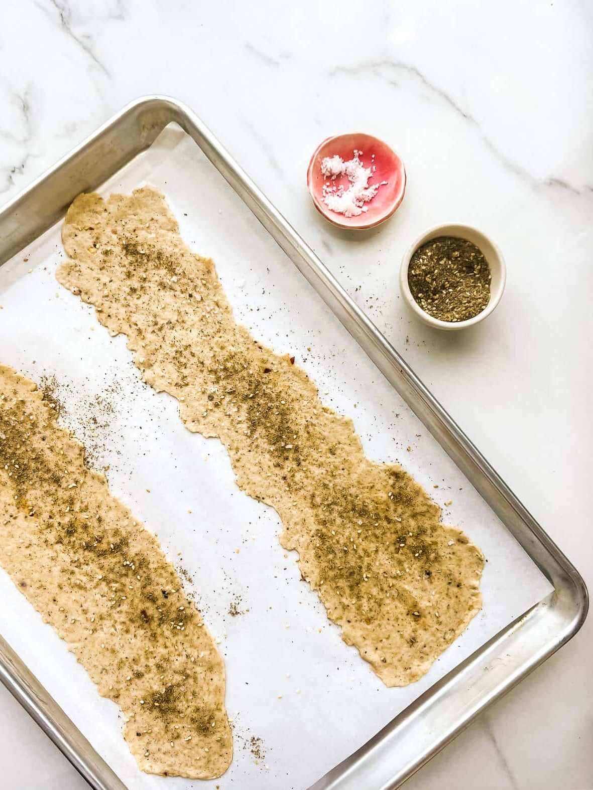 Sheets of rolled out cracker dough garnished with spices and salt, ready to be cut out and baked