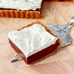 A square piece of a chocolate tart topped with whipped cream on a wooden board.