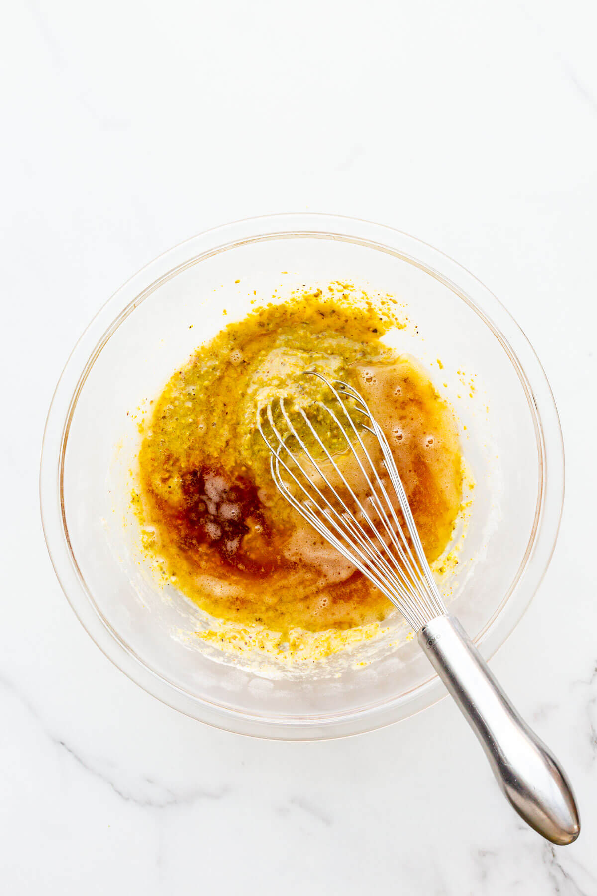 Adding brown butter to pistachio financier cake batter in a glass bowl with a whisk.