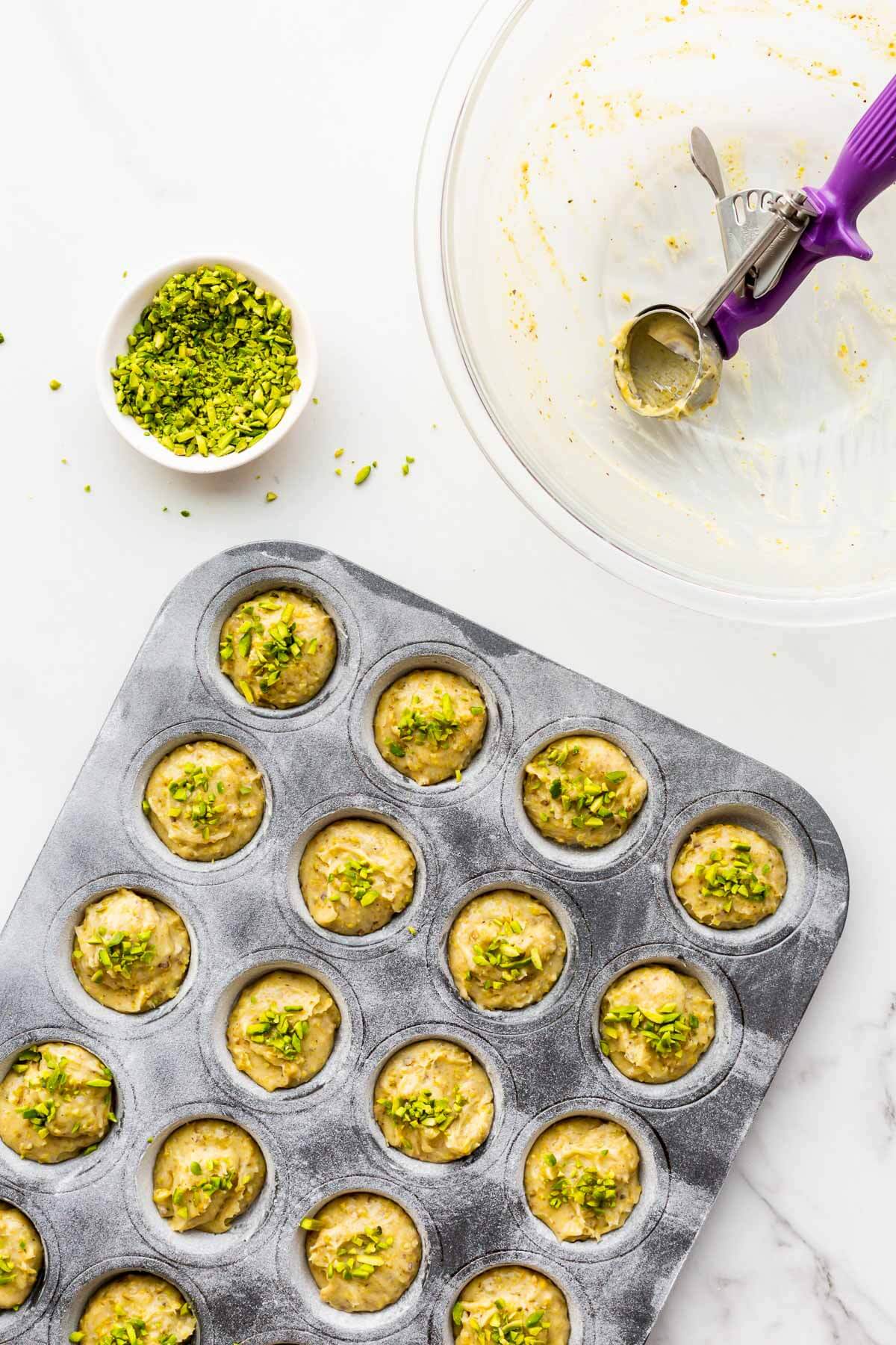 Scooping pistachio financier batter into a greased and floured mini muffin pan and sprinkling the tops with chopped pistachios before baking.