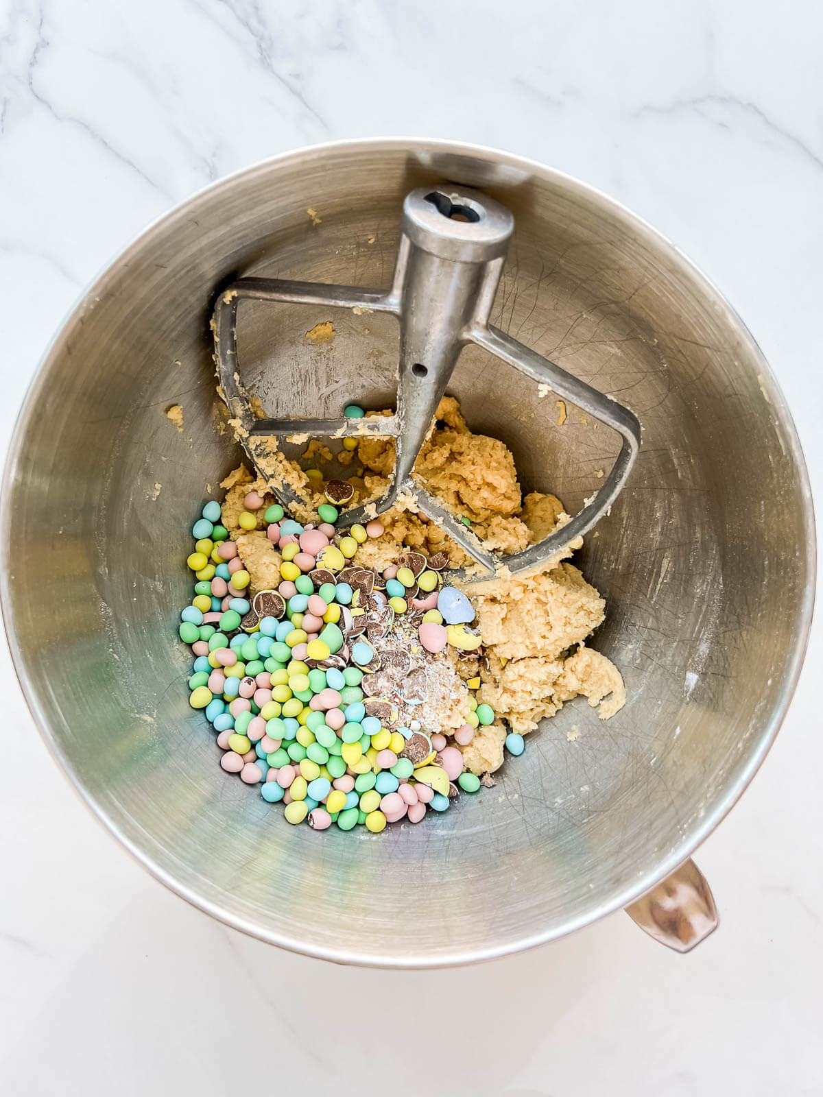 A mixer bowl with the paddle attachment with cookie dough topped with Easter Mini Eggs, ready to be mixed together.