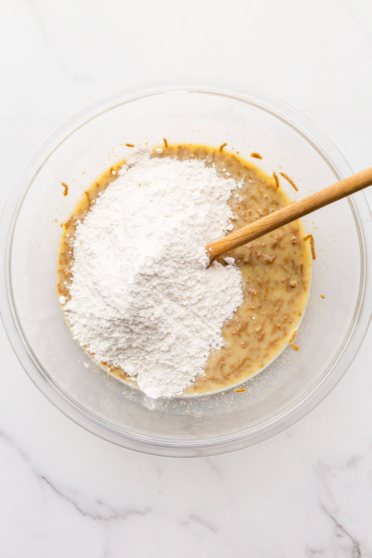 Stirring flour into bran muffin batter in a glass bowl with a wooden spoon.