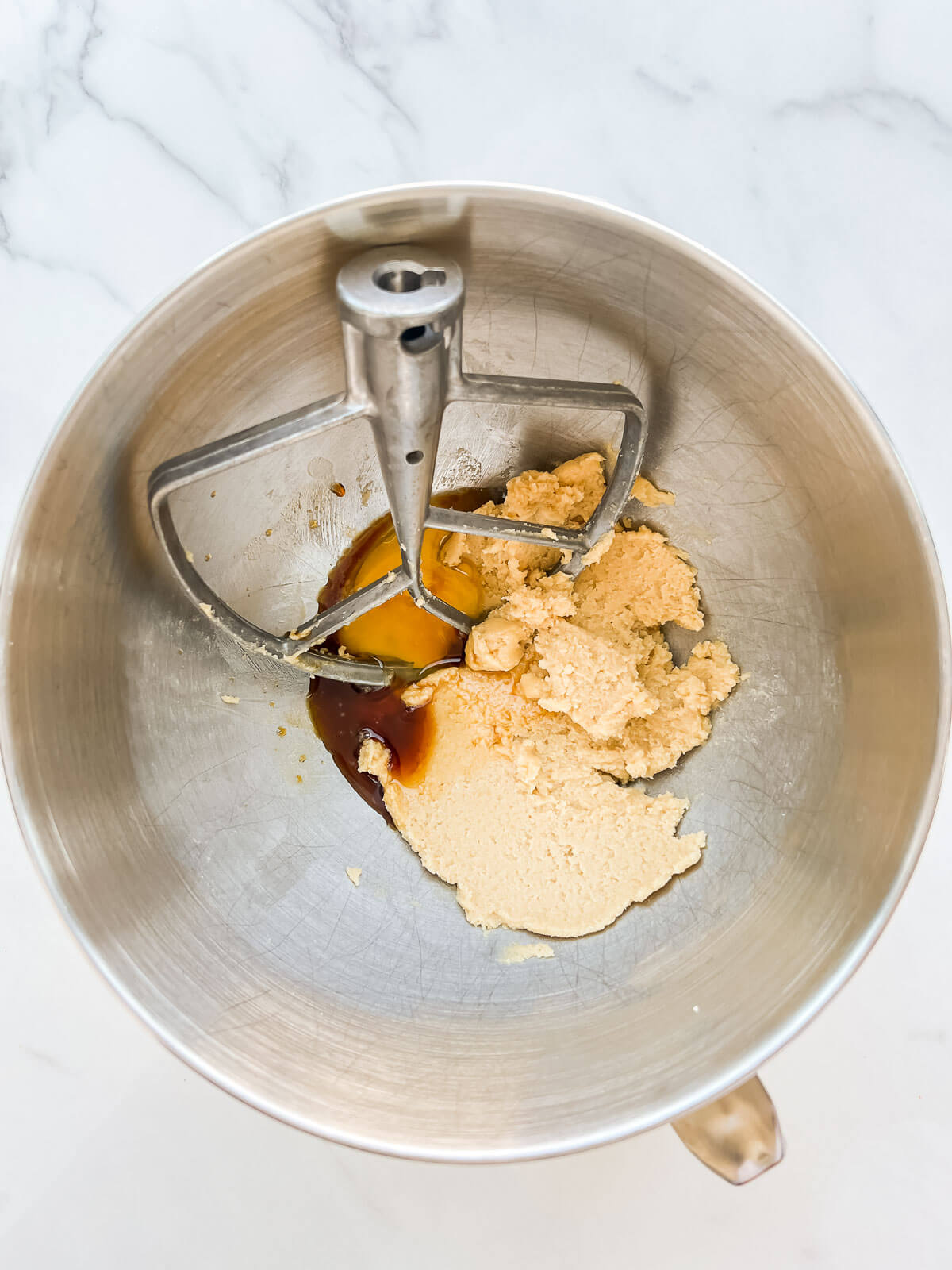 A mixer bowl with creamed butter and sugar, and the paddle attachment, with eggs and vanilla added to be mixed in next.