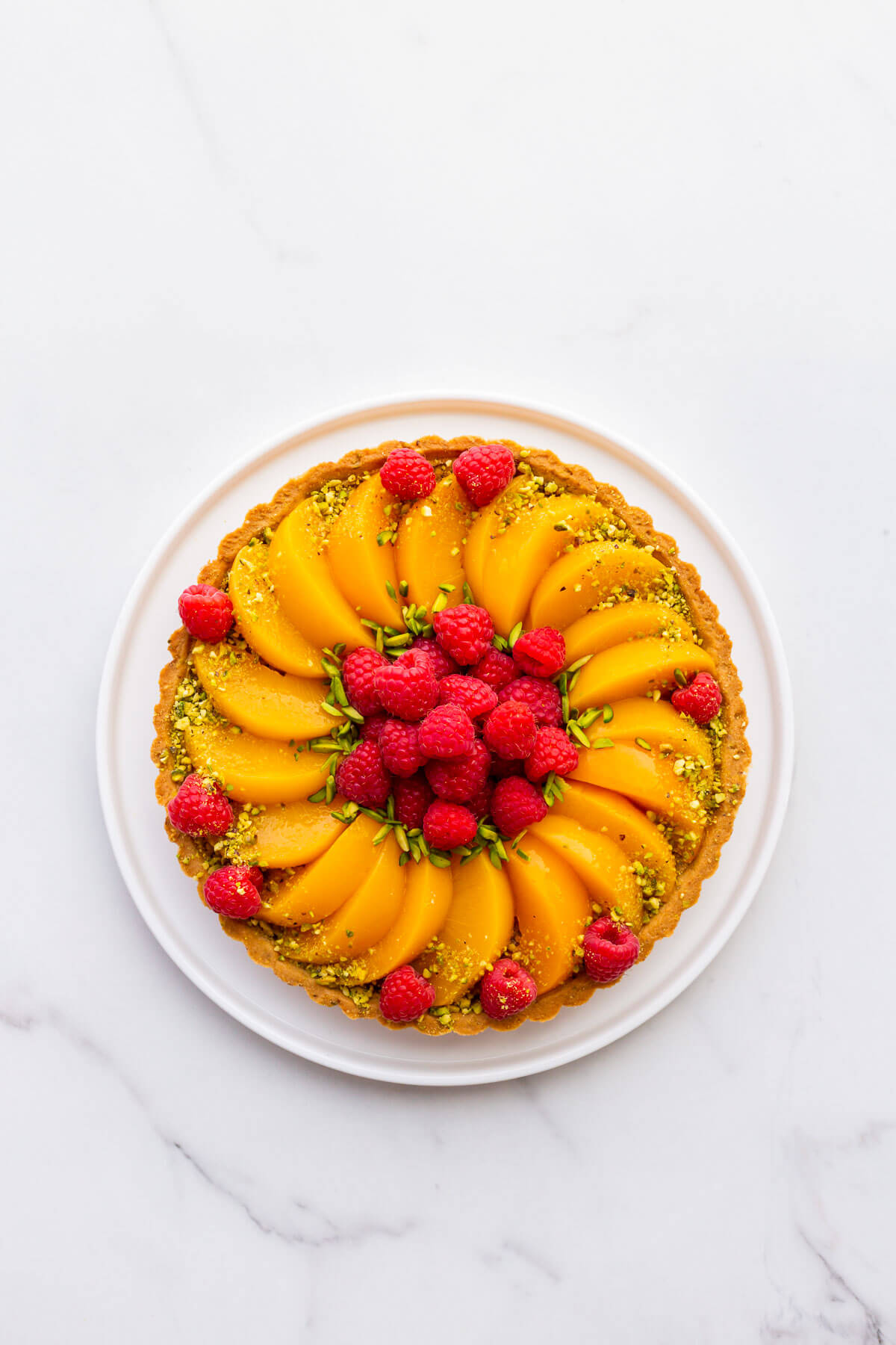 Pistachio tart topped with sliced peaches and fresh raspberries, and decorated with slivered and ground green pistachios.