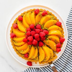 Slicing and serving a pistachio tart topped with sliced peaches and fresh raspberries, and decorated with slivered and ground green pistachios.