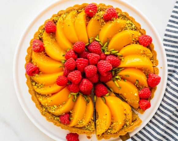 Slicing and serving a pistachio tart topped with sliced peaches and fresh raspberries, and decorated with slivered and ground green pistachios.