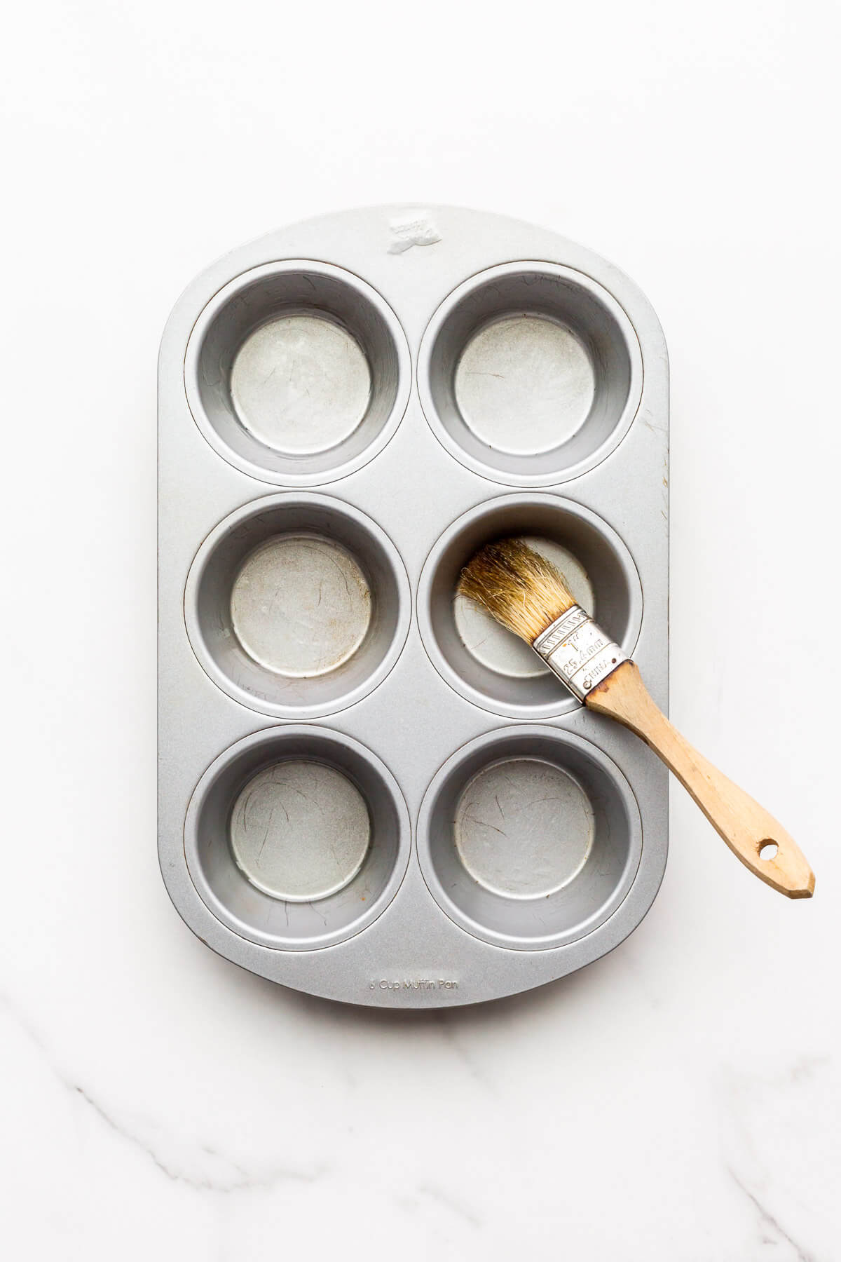 Brushing a muffin pan with canola oil to prevent homemade muffins from sticking to the pan when they bake.