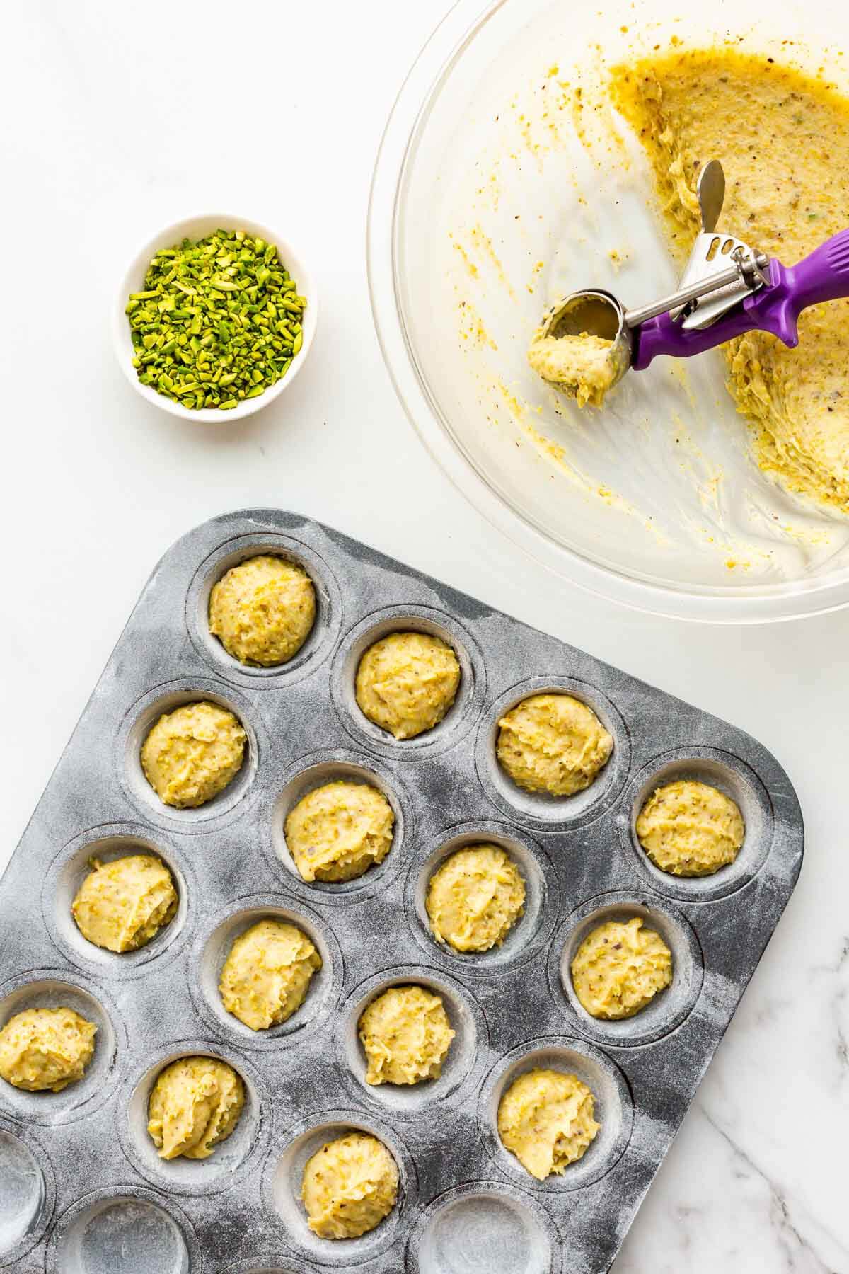 Scooping pistachio financier batter into a greased and floured mini muffin pan before sprinkling the tops with chopped pistachios before baking.