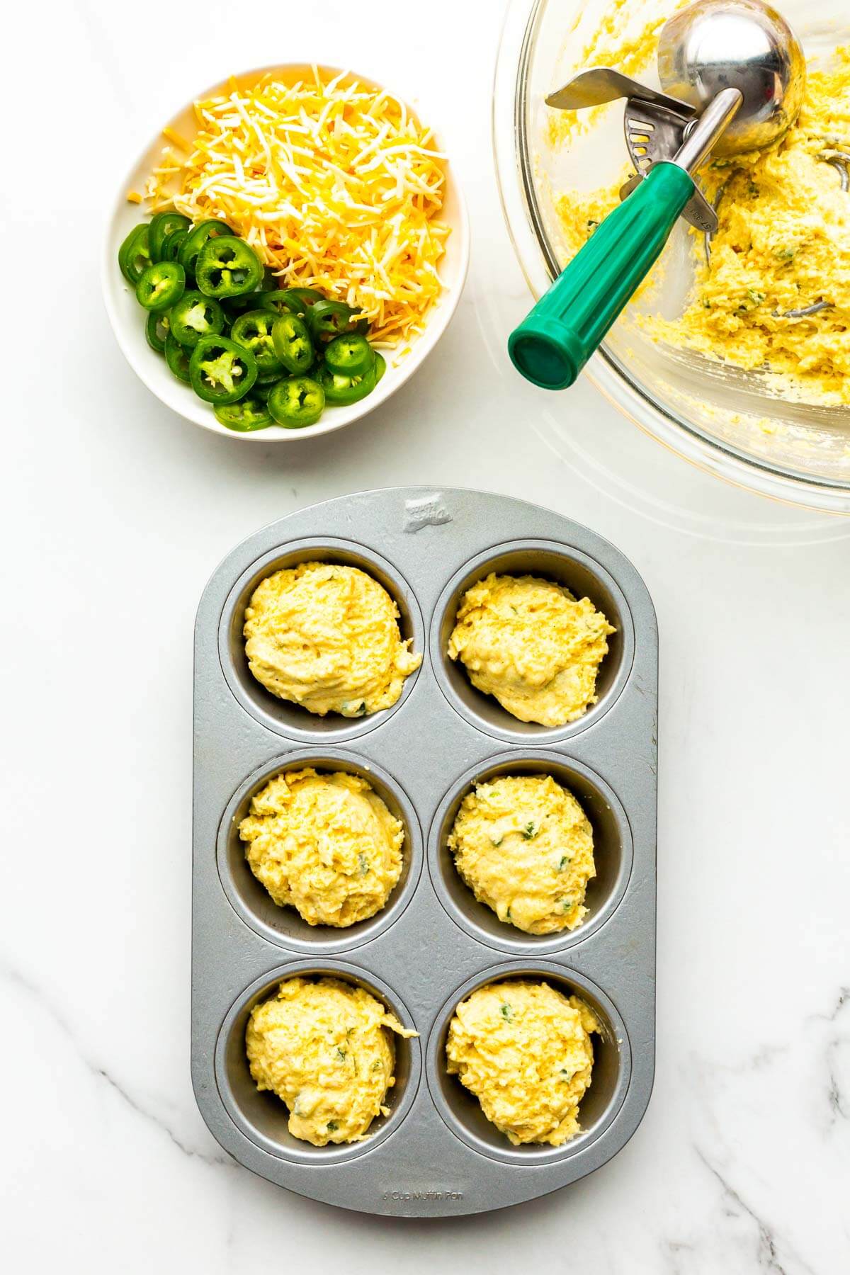 Scooping cornbread muffin batter into a greased 6-cup muffin pan.