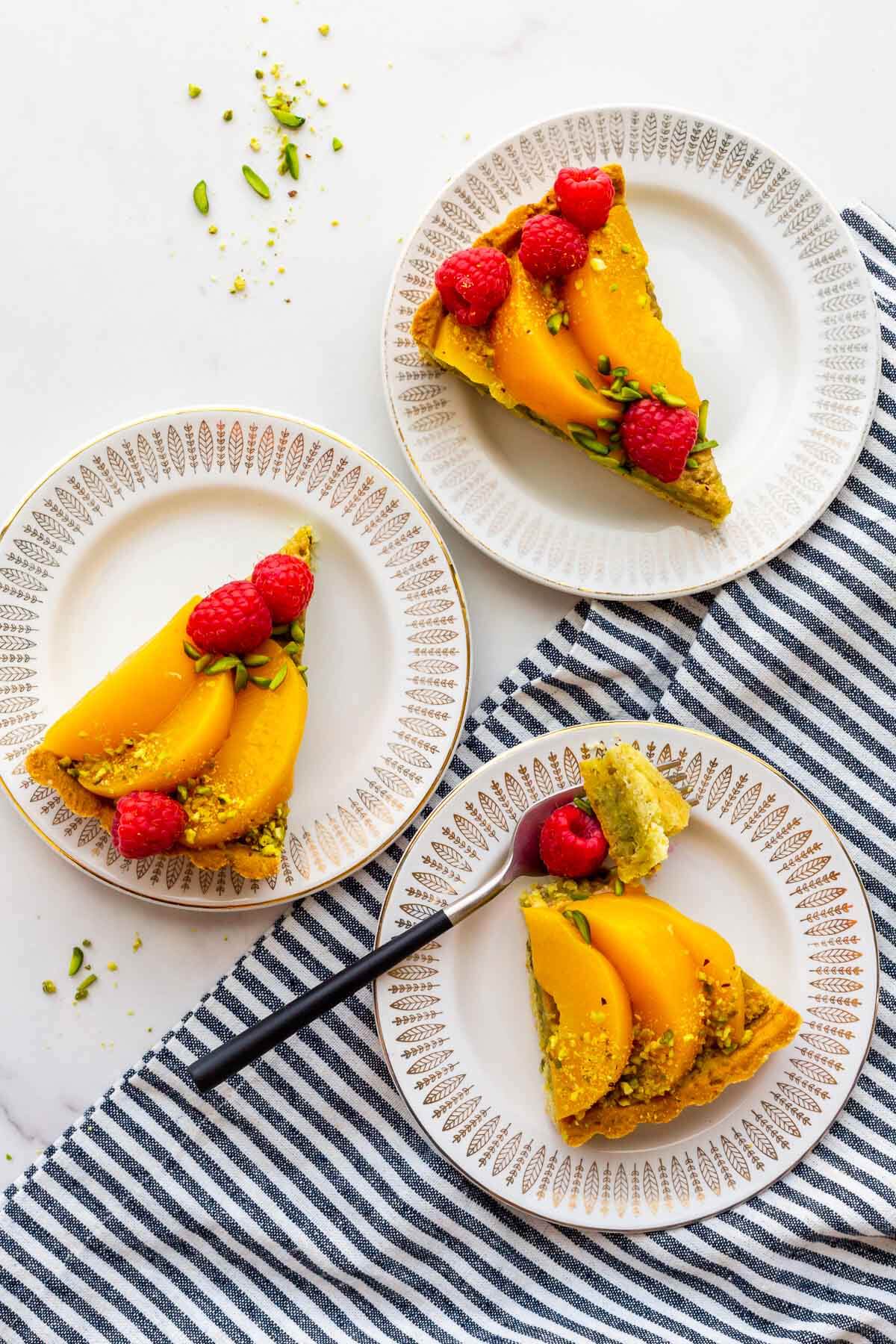 Three slices of pistachio tart with raspberries and peaches on plates.