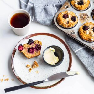 A plate with an apple blackberry muffin split open, served with butter and a cup of tea.