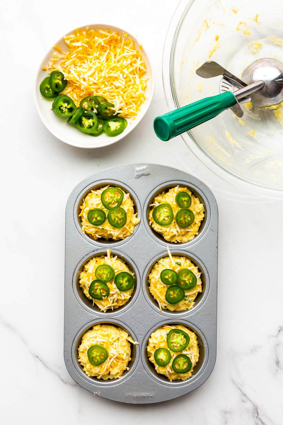 Scooping and garnishing cornbread muffin batter into a greased 6-cup muffin pan, topping with jalapeño and shredded cheddar.