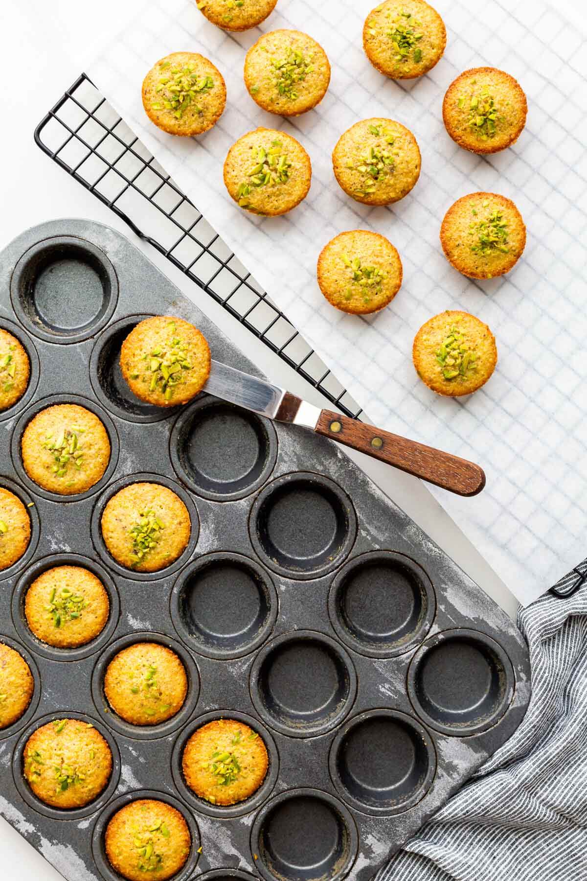 Transferring freshly baked pistachio financiers from a mini muffin pan to a cooling rack using a mini offset spatula to lift them.