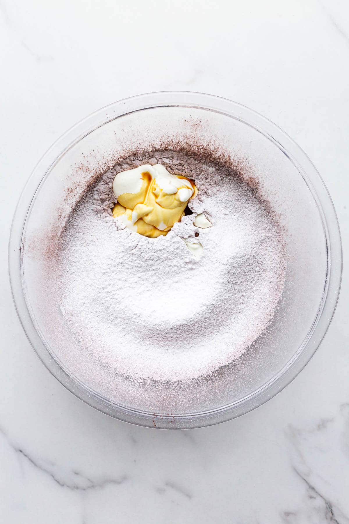 Adding sour cream and vanilla to a glass bowl with sifted icing sugar and cocoa powder to make a chocolate fudge frosting.