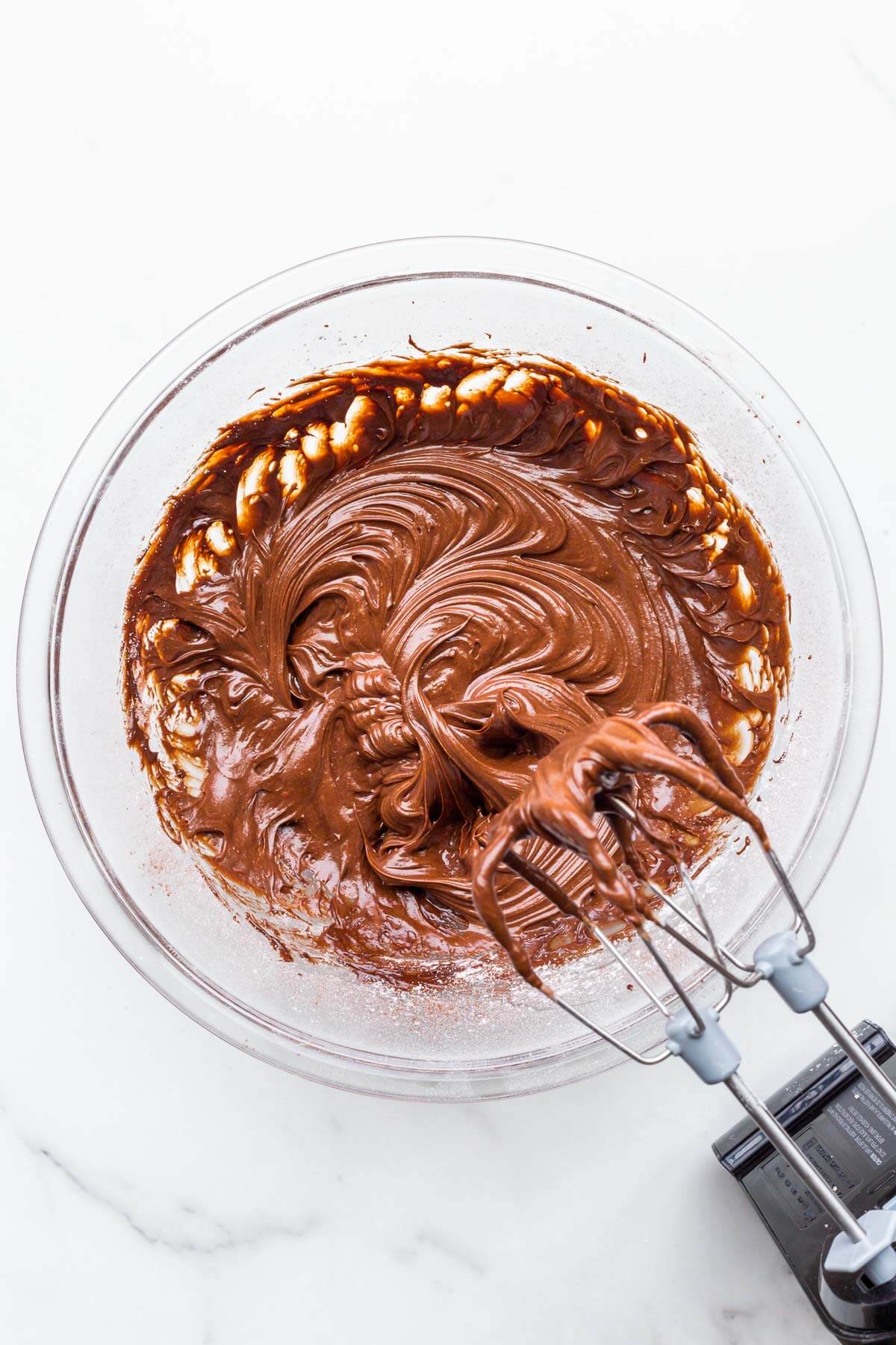 A glass bowl of glossy chocolate fudge frosting mixed with an electric mixer.