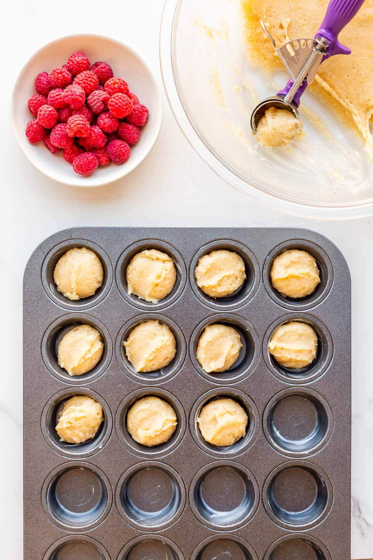 Scooping almond financiers batter into a mini muffin pan and garnishing each financier with a fresh raspberry before baking.