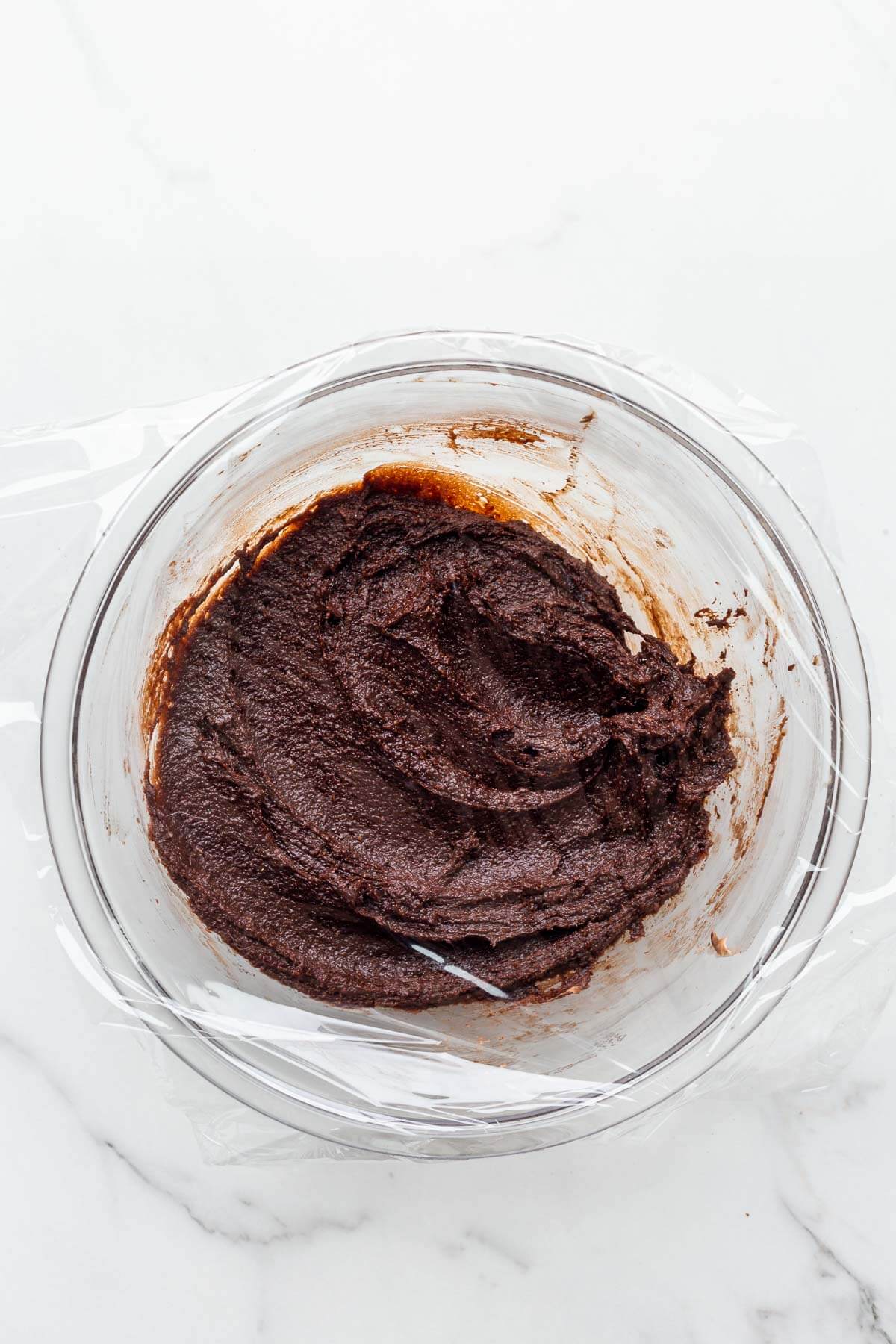 Chocolate financiers batter in a glass bowl wrapped in plastic wrap to chill it before baking.