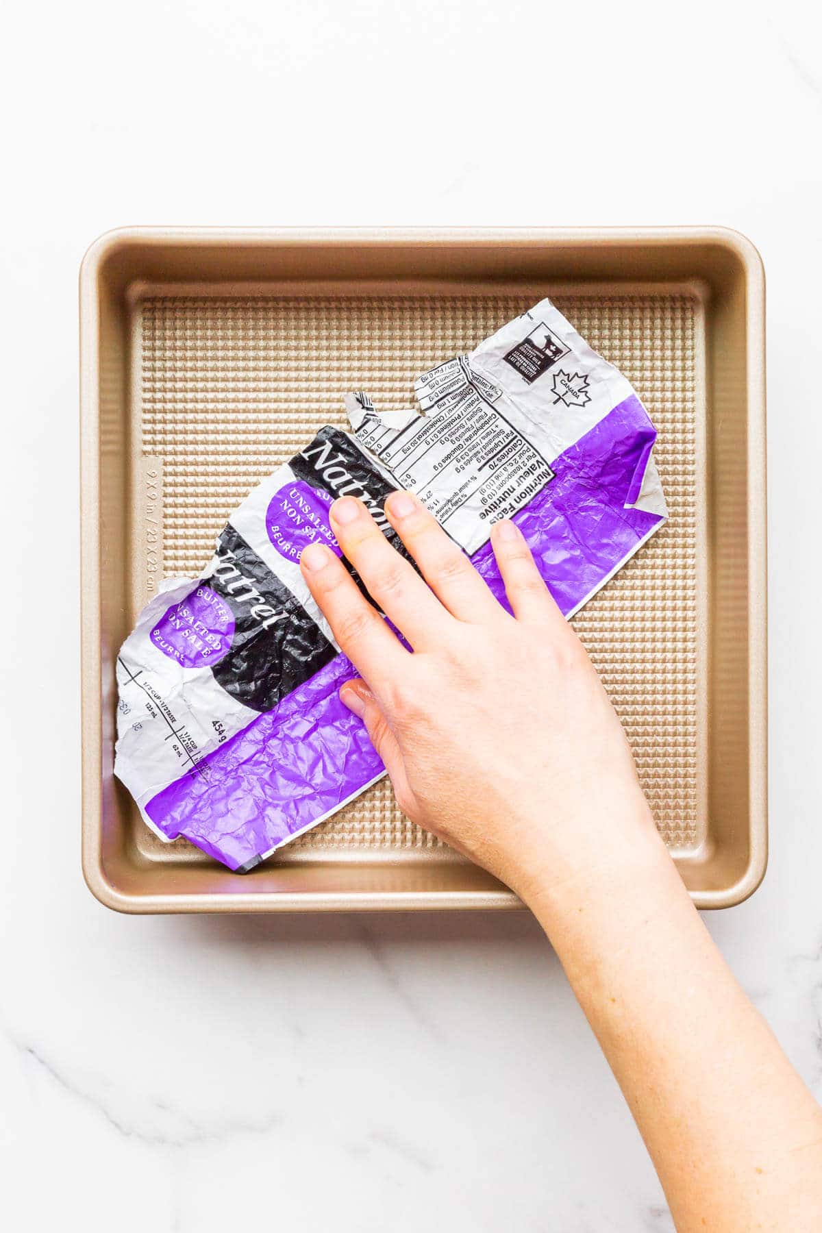 Buttering a 9x9 cake pan with softened butter and the help of the butter wrapper to smooth the butter over the entire surface of the pan.