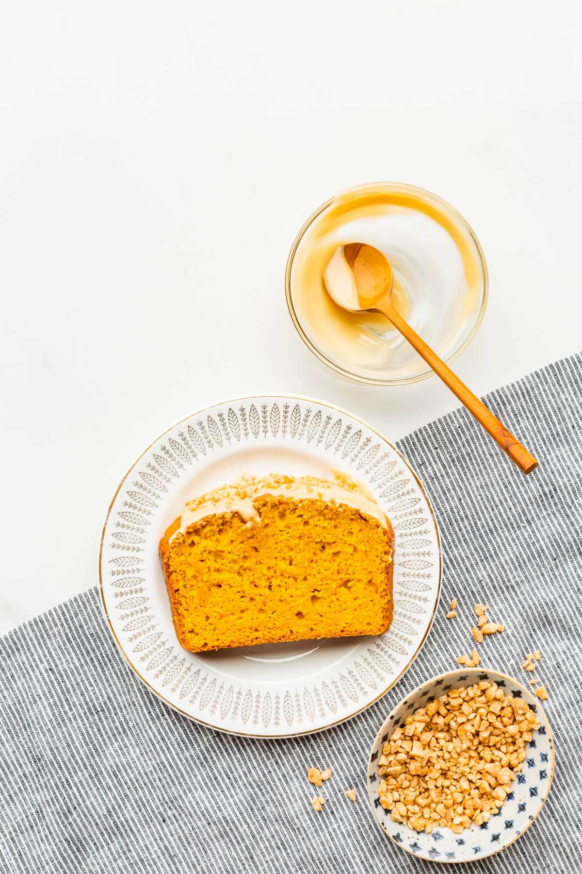 A slice of pumpkin bread on a dessert plate with small bowls of maple butter and maple flakes on the side.