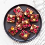 Chocolate brownies topped with raspberries and white chocolate chunks, cut into squares and served on a black plate.