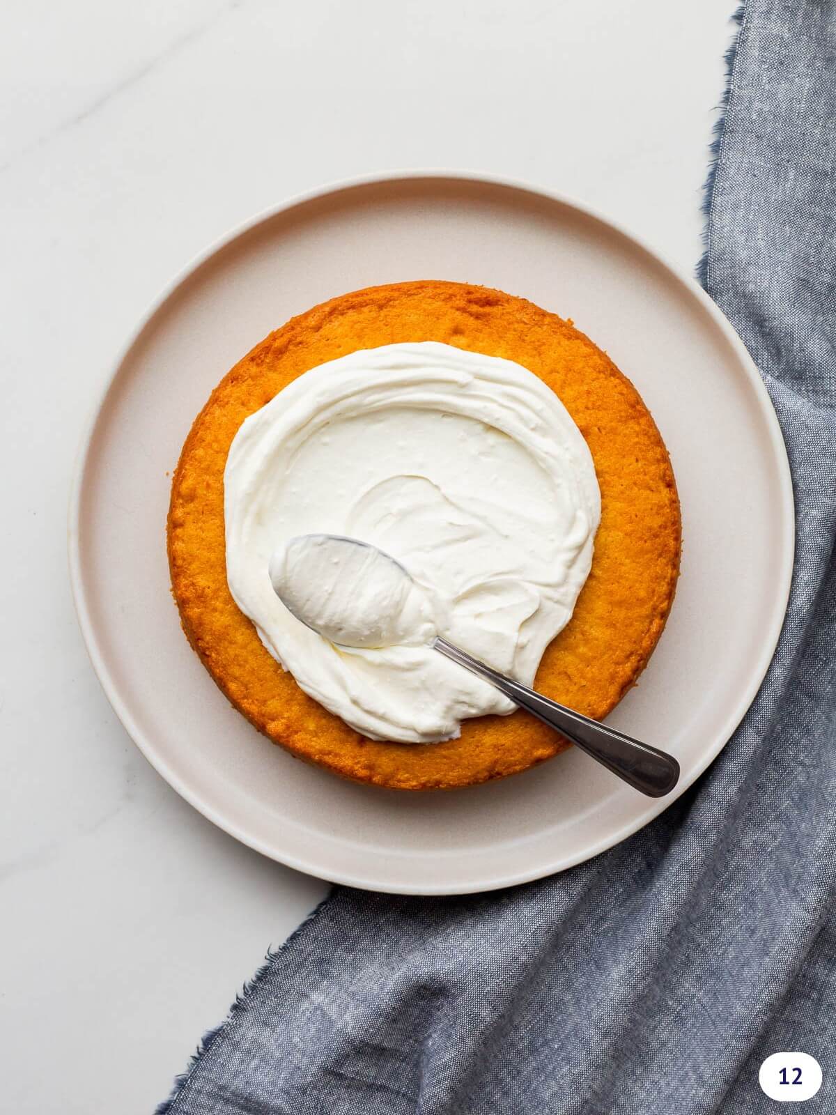 Smearing whipped cream on a single layer of cake set on a beige plate using a spoon to spread the cream.