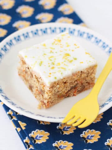 A square of carrot cake on a plate with a yellow fork.
