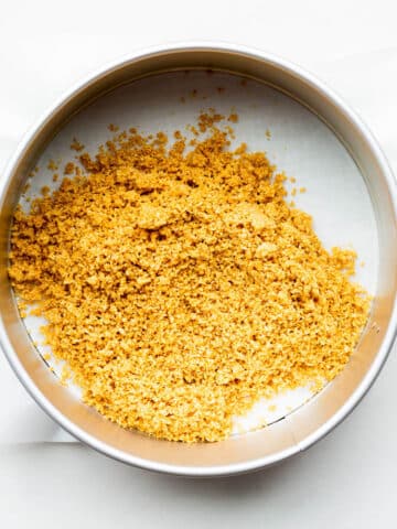 Graham cracker crumbs in a springform pan lined with parchment paper, to be spread out and pressed to form a crust for cheesecakes.