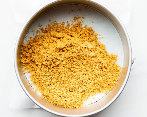 Graham cracker crumbs in a springform pan lined with parchment paper, to be spread out and pressed to form a crust for cheesecakes.