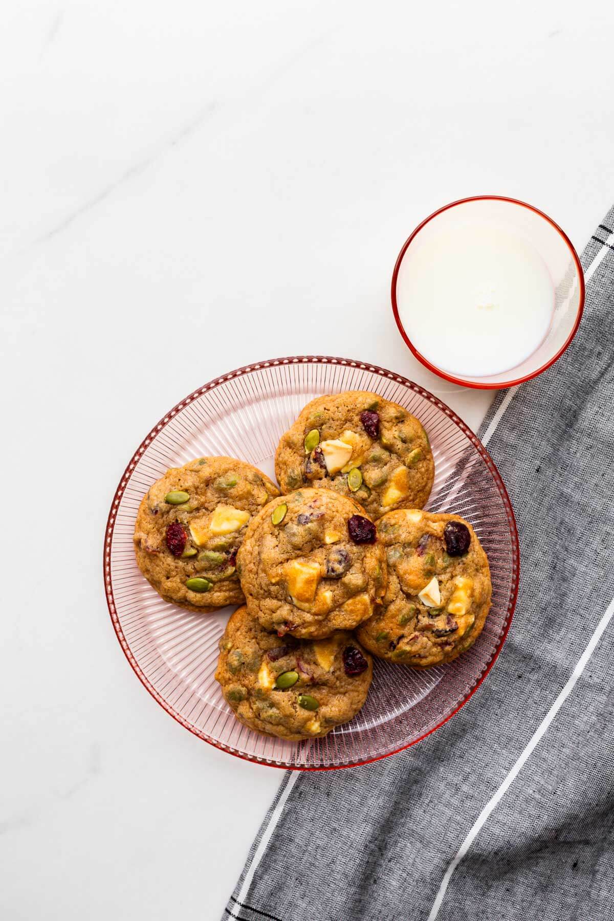 A plate of cookies featuring dried cranberries, white chocolate and pumpkin seeds with a glass of milk.