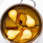 A saucepan with pears poached in a wine and spice-infused syrup with cinnamon sticks, star anise, vanilla bean, and orange peel.