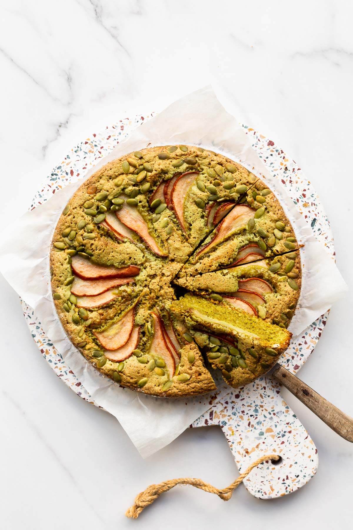 A moist pumpkin seed cake topped with thinly sliced pear being sliced and served. A slice is tilted slightly to show how vibrant green the crumb is.